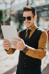 A man wearing a brown shirt, black vest, and sunglasses smiles while holding a tablet outdoors, conveying modern work, tech focus, and confident business lifestyle in an urban environment. © qunica.com