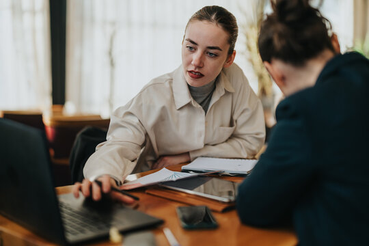 A group of young colleagues discuss a project at a round table, using laptops and notebooks. They collaborate, exchange ideas, and focus on planning and strategy in a modern office setting.