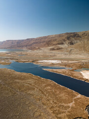 High-angle aerial view of geological sinkholes (collapse pits) forming along the rapidly receding shoreline of the Dead Sea. The image features contrasting colors of water-filled holes 