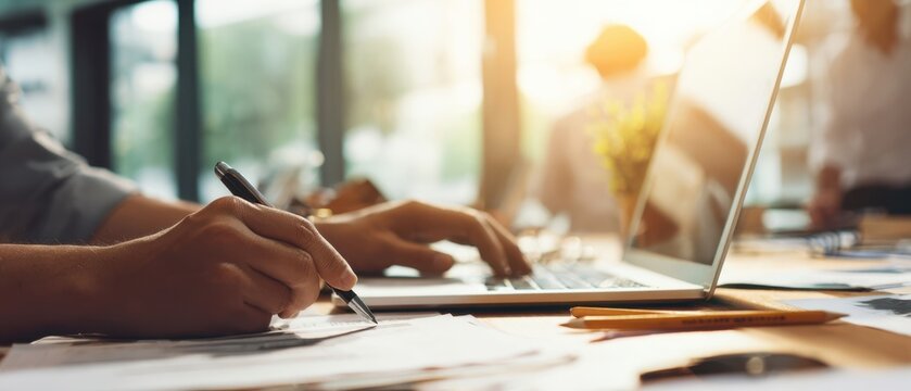 The laptop on a busy meeting table with hands writing and documents