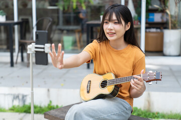 Young woman playing ukulele and recording video with smartphone outdoors