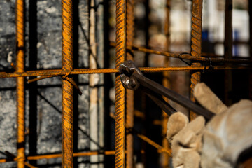 Close-up of a construction worker wearing protective gloves and using pliers to cut or tie steel rebar at a job site. Warm light highlights the texture of rusted metal and manual labor.