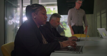 Focused professionals using laptops at meeting table while standing colleague observes discussion in collaborative corporate workspace