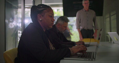 Focused professionals using laptops at meeting table while standing colleague observes discussion in collaborative corporate workspace