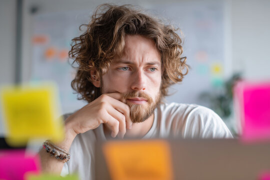 Pensive young man with curly hair and beard focusing on laptop amid colorful sticky notes — thoughtful portrait of remote work and creative planning