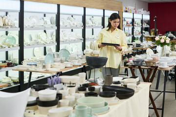 Young adult woman standing in tableware store holding digital tablet, checking inventory among shelves filled with plates, bowls, cups and various ceramic kitchenware items