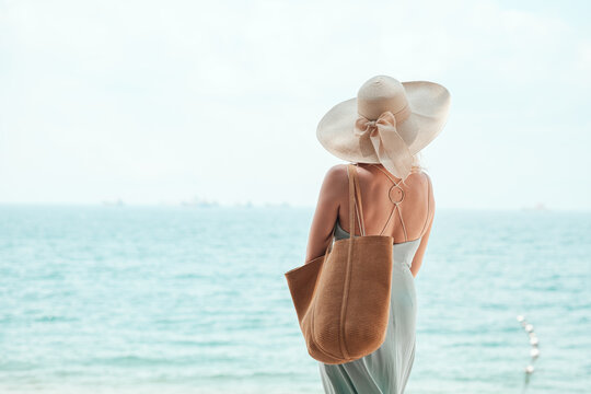 Elegant rear view of a young woman in a wide-brim sun hat gazing at the calm sea, evoking summer beach vacation vibes and serene coastal relaxation. Ocean horizon,  summer fashion