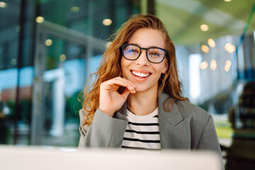 Close-up of business woman sitting at desk outdoors. Beautiful woman in stylish clothes enjoys working remotely on sunny day on terrace. Concepts of freelancing, modernity, style, and business.