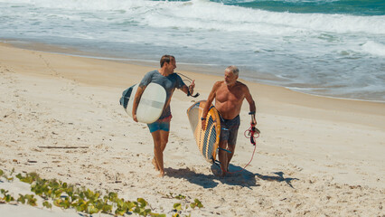 Young and elderly surfers walk with surfing boards on the beach in Brazil