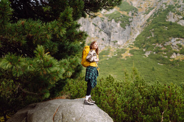 Obraz premium A female hiker with a backpack stands against a mountain backdrop at sunset. A happy woman enjoys the mountains and cliffs on her journey. Adventure, trail, and active lifestyle concepts.