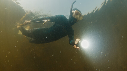 River spearfishing. Woman with speargun and torch slowly swims underwater in the murky river and explores area full of underwater plants