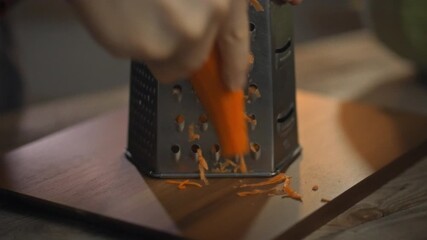 A person is grating fresh carrots on a box grater, focusing on preparing healthy food in a cozy kitchen. The kitchen has a warm ambiance, highlighting the care in meal preparation