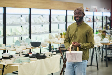 Black young adult man standing in kitchenware store holding shopping basket, smiling while browsing tableware selection, shelves filled with plates and bowls in background