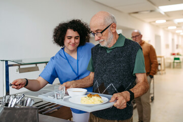 Senior man having lunch with supportive caregiver in community center cafeteria.