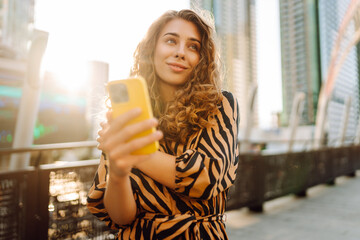 A cheerful woman poses on the street with a phone against the backdrop of a bustling metropolis. A...