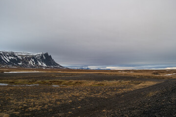 Die dunklen Klippen der Halbinsel Snæfellsnes erheben sich imposant aus der flachen Ebene. Der graue, eintönige Wolkenschleier hängt tief und ist fast zum Greifen nah.