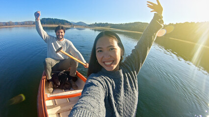 A joyful couple paddles a canoe on a serene lake, capturing a selfie. The woman smiles broadly, raising her arms in excitement, while the man holds the paddle.