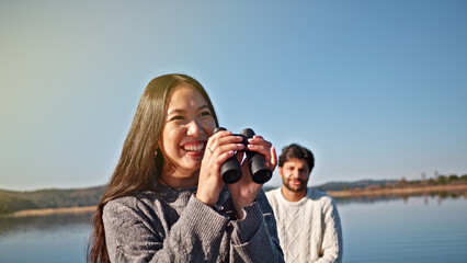 A young woman with long dark hair, wearing a gray sweater, smiles while using binoculars by a serene lake. A man stands behind her, suggesting a leisurely outing.