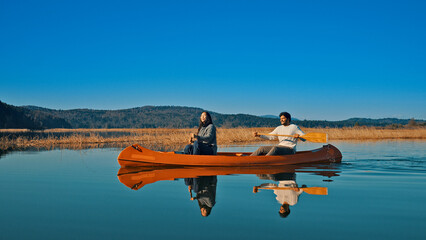 Two individuals paddle a canoe on a serene lake, surrounded by autumnal reeds and distant hills....