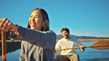 A joyful Asian woman paddles a canoe, smiling, while her partner rows. They enjoy a serene outdoor adventure, embodying a healthy, active lifestyle.