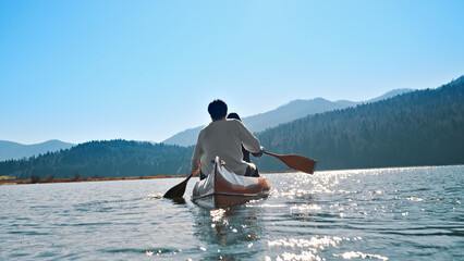 A couple paddles a canoe on a serene lake, surrounded by majestic mountains. The clear blue sky and calm waters reflect a peaceful, active lifestyle, ideal for promoting outdoor adventures.