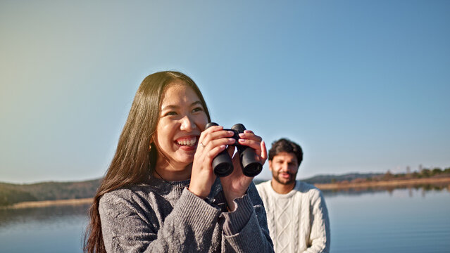 A young woman with long dark hair, wearing a gray sweater, smiles while using binoculars by a serene lake. A man stands behind her, suggesting a leisurely outing.