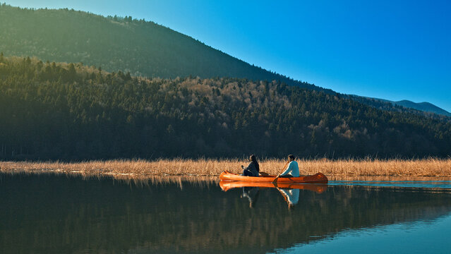 A couple paddles a canoe on a serene lake, surrounded by forested hills. The calm water reflects the clear blue sky, capturing a peaceful moment of connection in nature. - Powered by Adobe