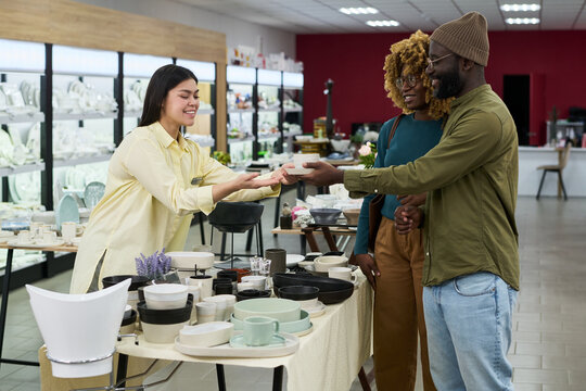 Young Caucasian woman handing ceramic bowl to smiling Black man and Black woman standing together at pottery shop counter, customers interacting with shop assistant during purchase