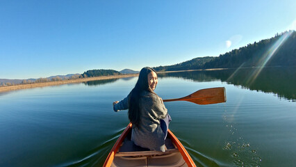 A woman paddles a canoe on a serene lake, surrounded by lush hills under a clear blue sky. The sun casts a gentle glow, highlighting her joyful expression and the tranquil waters.