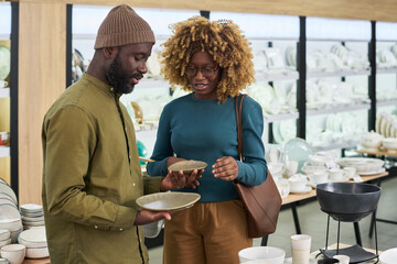 Black man and Black woman shopping together in store, examining ceramic plates, standing side by side, both appearing as young adults, shelves with tableware in background