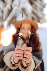 A cheerful woman in a hat in a snow-covered park enjoys sparklers. A beautiful woman in warm...