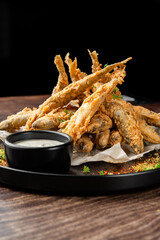 Crispy deep-fried small fish served with a creamy dipping sauce on a black plate with parchment paper and herbs.