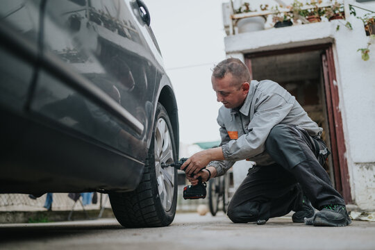 A mechanic works on a car tire in an outdoor setting, kneeling beside the front wheel and using a cordless drill. This image conveys automotive repair, maintenance, and roadside service.