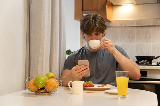 Drinking coffee and checking phone caucasian young man sitting at kitchen table during morning breakfast enjoying warm drink routine. Healthy Breakfast. Home Kitchen Lifestyle