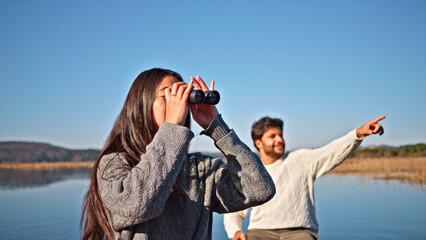 A young couple enjoys a serene lakeside outing. The woman, with long dark hair, uses binoculars to scan the horizon, while the man, dressed in a white sweater.