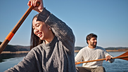 A joyful couple paddles a canoe on a serene lake, embracing a weekend adventure. The woman, in a cozy sweater, raises her paddle with a smile, while her partner rows behind her.
