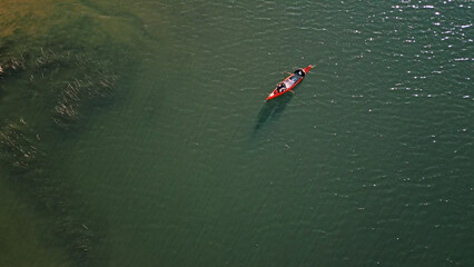 A drone shot captures a couple paddling a vibrant red canoe on a serene, sunlit lake, embodying weekend adventure and active lifestyle. The tranquil waters and lush greenery reflect.