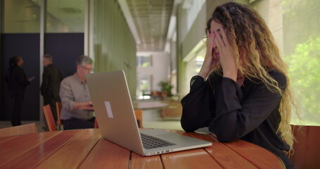 Obraz premium Frustrated woman leaning back and pressing face with hands while sitting at laptop in modern office lounge, overwhelmed expression indicating work stress