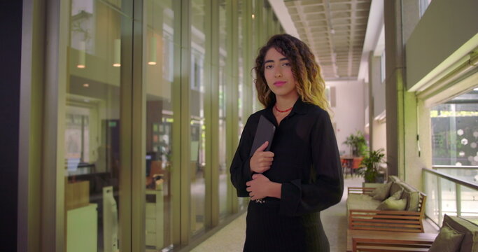 Full body portrait of young woman holding tablet, standing in modern office corridor with confident stance, facing forward in professional workspace