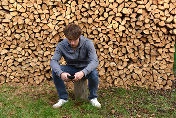 Young man sitting with phone by firewood stack