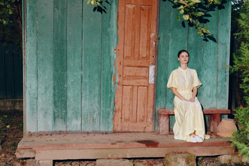 Woman sits on a wooden bench outside a rustic green wall beside a weathered doorway, wearing a pale yellow dress. The scene exudes calm vintage charm and quiet contemplation in a simple outdoor