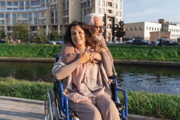 Senior woman in wheelchair walking with caregiver old man on road in park. Elderly family couple...