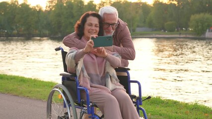 Senior couple woman in wheelchair caregiver old man making selfie enjoying walking. Elderly family man supporting embracing woman in chair for people with disability taking photo. Rehabilitation