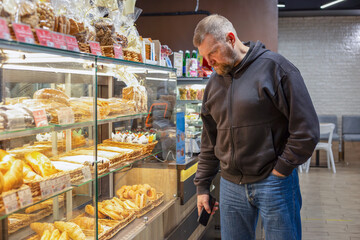 A man in a cafe is choosing pastries near the counter. Behind the glass, there are croissants, filled rolls, doughnuts, muffins, cookies in baskets. Indoor bakery setting, food choice, daily routine.
