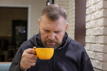 Man sitting in a cozy cafe holding a yellow coffee cup, focusing on the warm drink in his hands. Mature man enjoying his coffee break. Close-up on the face and hand holding a warm orange cup.