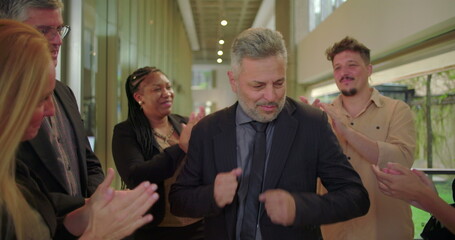 Grateful male professional smiling while receiving applause from supportive colleagues in office hallway, emotional moment of recognition and appreciation