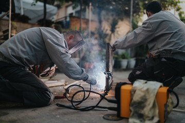 Two workers in protective gear weld a metal frame outdoors, sparks and smoke rise while they secure the joint. Nearby tools and cables create a makeshift workshop setting.