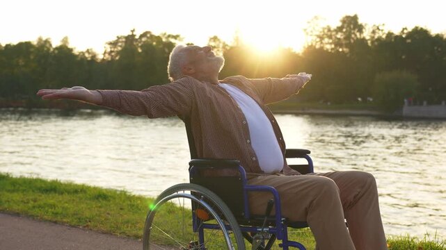 Happy senior man in wheelchair raised hands enjoying freedom walking on road in city park outdoor. Old happy man in chair for people with disability has a hope. Disability disease rehabilitation