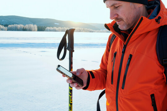 Middle aged Caucasian man standing outdoors on snowy landscape holding ski poles and using smartphone, wearing winter clothing and looking at device with focused expression