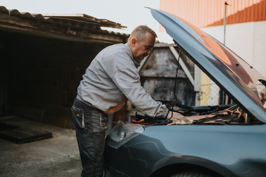 A skilled mechanic leans over the engine, checking cables and components under the hood of a blue car in an outdoor storage area for maintenance and repair.